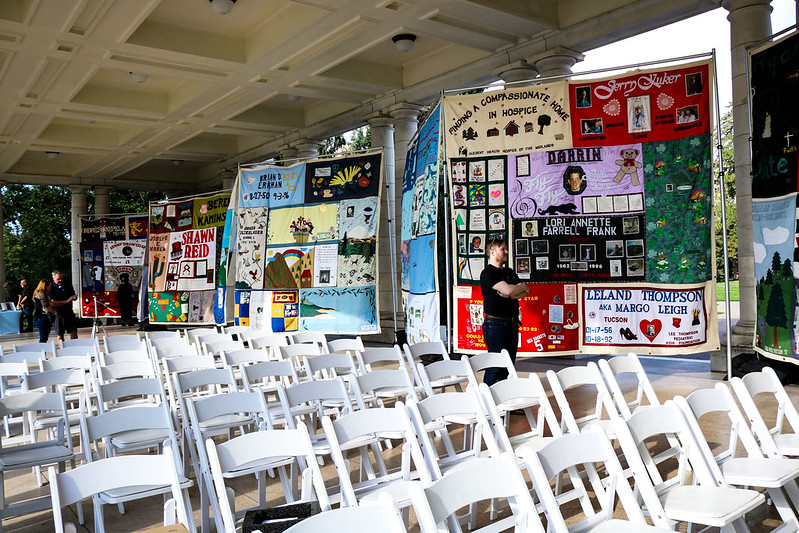 An image of empty rows of white chairs, with 8 panels of the AIDS Quilt hanging in the background.